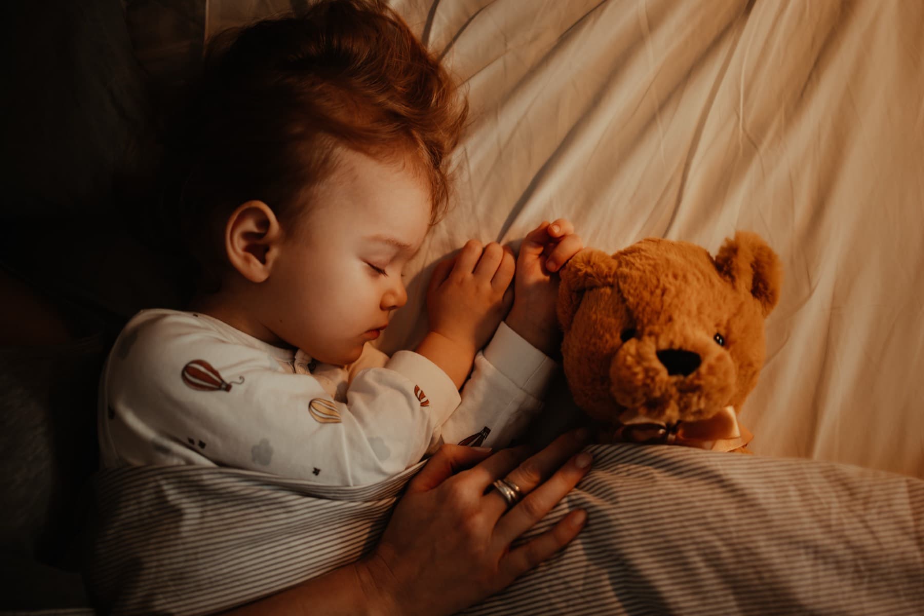 Slapend kindje in bed met een knuffelbeer, met de hand van een ouder op de deken